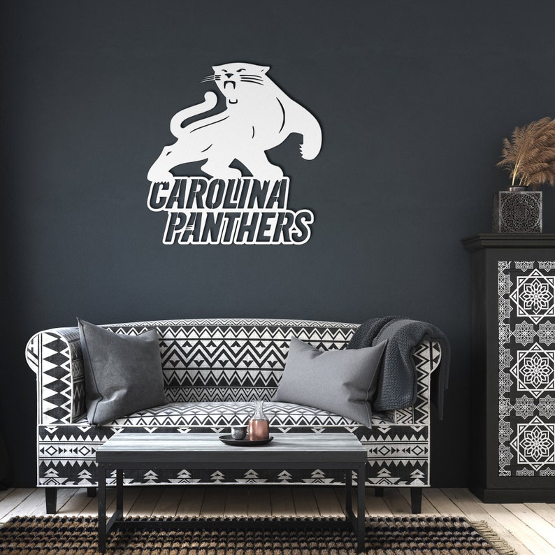 Puede incluir: Logotipo blanco de los Carolina Panthers, con un felino y el nombre del equipo, en una pared oscura. El logotipo est&aacute; encima de un sof&aacute; con un patr&oacute;n geom&eacute;trico en blanco y negro, una mesa de centro y una alfombra.