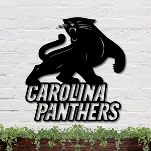 Puede incluir: Letrero met&aacute;lico negro de los Carolina Panthers con una silueta de pantera rugiente. El nombre del equipo se muestra en letras blancas en negrita debajo de la pantera. El letrero est&aacute; montado en una pared de ladrillo blanco.