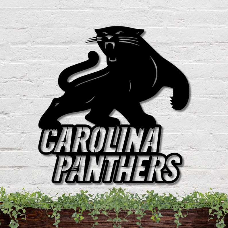 Puede incluir: Letrero met&aacute;lico negro de los Carolina Panthers con una silueta de pantera rugiente. El nombre del equipo se muestra en letras blancas en negrita debajo de la pantera. El letrero est&aacute; montado en una pared de ladrillo blanco.