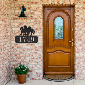 May include: Black metal house number sign with three horses running in front of the numbers "1749". The sign is mounted on a brick wall next to a wooden door.