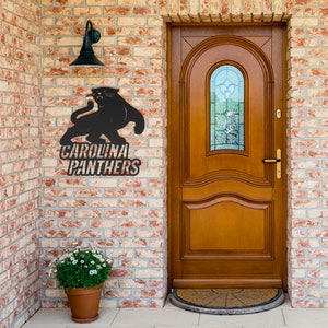 Puede incluir: Un letrero met&aacute;lico negro con el logotipo de los Carolina Panthers, montado en una pared de ladrillo. El letrero representa una pantera y las palabras "Carolina Panthers". Una l&aacute;mpara exterior negra est&aacute; encima del letrero. Una puerta marr&oacute;n est&aacute; a la derecha.