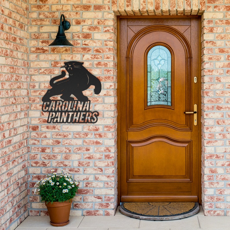 Puede incluir: Un letrero met&aacute;lico negro con el logotipo de los Carolina Panthers, montado en una pared de ladrillo. El letrero representa una pantera y las palabras "Carolina Panthers". Una l&aacute;mpara exterior negra est&aacute; encima del letrero. Una puerta marr&oacute;n est&aacute; a la derecha.
