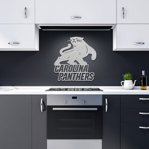 Puede incluir: Un logotipo plateado de los Carolina Panthers montado en una pared gris oscuro sobre una estufa. El logotipo presenta una pantera con el nombre del equipo en letras en negrita. La cocina tiene gabinetes blancos y un horno negro.