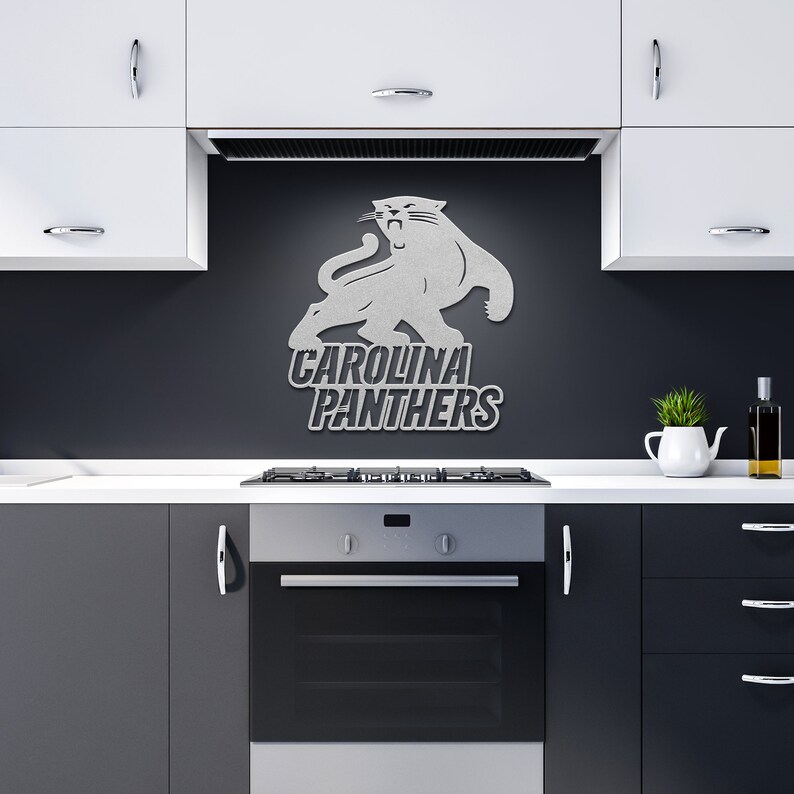 Puede incluir: Un logotipo plateado de los Carolina Panthers montado en una pared gris oscuro sobre una estufa. El logotipo presenta una pantera con el nombre del equipo en letras en negrita. La cocina tiene gabinetes blancos y un horno negro.