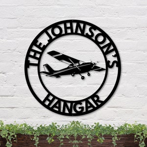 May include: Black metal circle sign with the text "The Johnsons Hangar" and a silhouette of a small airplane inside the circle.