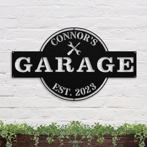 May include: Black metal garage sign with white lettering. The sign reads "Connor's Garage Est. 2023" with a pair of crossed wrenches in the center.