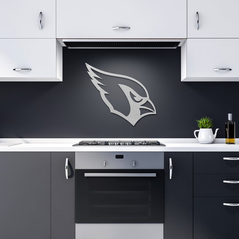 May include: A silver metal wall decor featuring the Arizona Cardinals logo. The logo is a stylized bird's head with a red beak and a white eye. The decor is mounted on a black wall in a kitchen setting.