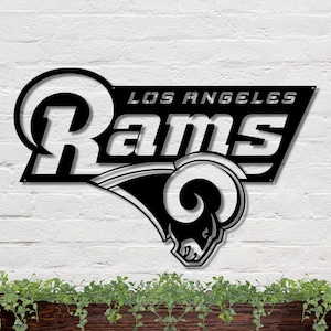 May include: Black metal wall art featuring the Los Angeles Rams logo and the words "LOS ANGELES Rams". The design includes the team's ram head logo. The artwork is mounted on a white brick wall.