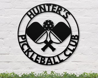 Może przedstawiać: Czarny metalowy znak z tekstem "Hunter's Pickleball Club" w kółku. Znak przedstawia dwie skrzyżowane rakiety do pickleballa i piłkę do pickleballa.