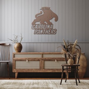 Puede incluir: Una pieza de arte mural de metal marr&oacute;n con el logotipo de los Carolina Panthers, un felino gru&ntilde;endo sobre el nombre del equipo. La obra de arte est&aacute; montada en una pared gris con paneles, encima de un mueble de madera con detalles tejidos. La habitaci&oacute;n est&aacute; decorada con plantas secas y una alfombra.