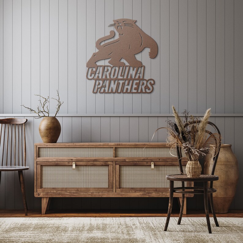 Puede incluir: Una pieza de arte mural de metal marr&oacute;n con el logotipo de los Carolina Panthers, un felino gru&ntilde;endo sobre el nombre del equipo. La obra de arte est&aacute; montada en una pared gris con paneles, encima de un mueble de madera con detalles tejidos. La habitaci&oacute;n est&aacute; decorada con plantas secas y una alfombra.