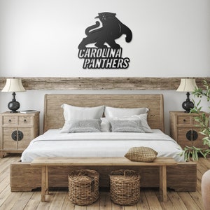 Puede incluir: Arte mural con el logotipo de los Carolina Panthers en metal negro. El logotipo presenta una silueta de pantera sobre las palabras "Carolina Panthers" en negrita y blanco. La obra de arte est&aacute; montada en una pared blanca sobre una estructura de cama de madera.