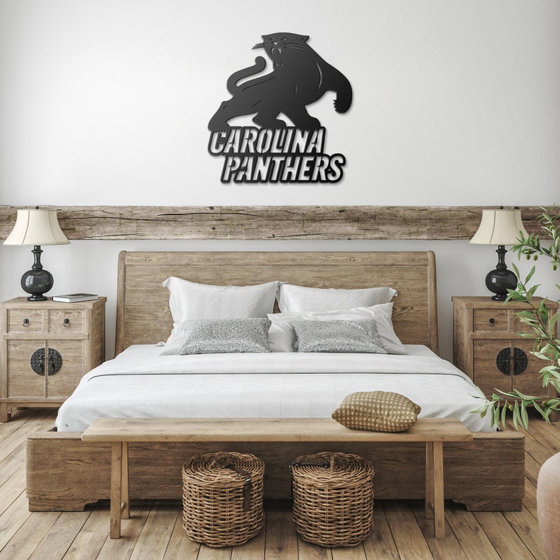 Puede incluir: Arte mural con el logotipo de los Carolina Panthers en metal negro. El logotipo presenta una silueta de pantera sobre las palabras "Carolina Panthers" en negrita y blanco. La obra de arte est&aacute; montada en una pared blanca sobre una estructura de cama de madera.