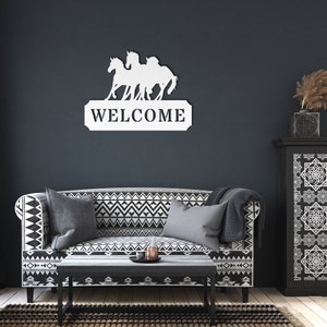 May include: White metal sign with three horses running in front of the word "WELCOME". The sign is mounted on a dark blue wall above a black and white patterned sofa.