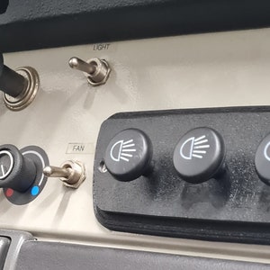 May include: Close-up of a car's dashboard with a row of black toggle switches and a panel of four round black buttons labeled with white icons of headlights. The panel is labeled "FAN" and "LIGHT".