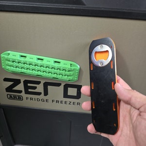 Puede incluir: Un abridor de botellas negro y naranja con un abridor de tapas plateado. El abridor de botellas está unido a un panel negro y naranja con una superficie texturizada. El panel tiene cuatro ranuras en el lateral. El texto "ZERO" y "ARB FRIDGE FREEZER" es visible en la superficie detrás del abridor de botellas.