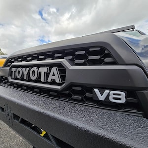 May include: Close-up of a black Toyota truck grille with the brand name in silver lettering. The grille features a honeycomb pattern and the letters "V8" in silver. The bumper is black and textured.