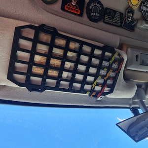 May include: Black metal grid organizer for a car's sun visor. The grid has a black and yellow elastic strap attached to it. The grid is mounted to the sun visor with two black clips.