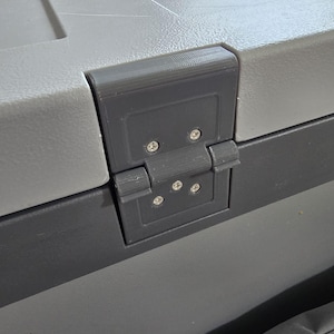 May include: Close-up of a gray and dark gray plastic hinge with screws. The hinge is attached to a gray and dark gray plastic container. The hinge is rectangular and has a series of small screws holding it together.