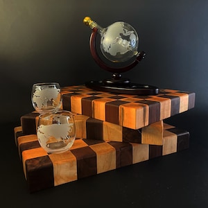 May include: A set of three clear glass tumblers with etched world maps, a globe-shaped decanter with a wooden stand, and a wooden cutting board with a checkerboard pattern.