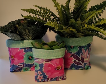 May include: Three fabric plant pots with floral patterns in shades of pink, blue, and green. The pots are filled with green plants, including a fern and a succulent.