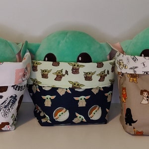 May include: Three green plush Baby Yoda toys are sitting inside fabric storage bins. Each bin has a different Star Wars themed fabric pattern. The first bin has a white fabric with a black and white Star Wars character pattern. The second bin has a green fabric with a black and white Baby Yoda pattern. The third bin has a brown fabric with a black and white Star Wars character pattern.