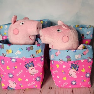 Może przedstawiać: Trzy różowe i niebieskie pojemniki do przechowywania z motywem Peppa Pig. Pojemniki mają niebieską i białą tkaninę w wzory z postaciami Peppa Pig na górze i różową tkaninę z postaciami Peppa Pig na dole. Pojemniki są wypełnione różowymi pluszowymi zabawkami Peppa Pig.