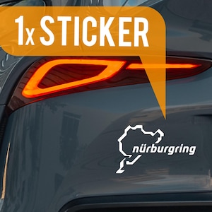 Op de afbeelding: Een witte sticker met de omtrek van het Nürburgring-circuit en tekst, bevestigd aan de achterkant van een grijze auto. De sticker bevindt zich onder het achterlicht. De afbeelding bevat de tekst "1x STICKER".