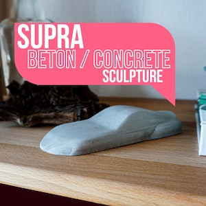 Könnte beinhalten: Graue Beton-Skulptur eines Autos auf einem Holzregal. Die Skulptur ist eine Miniatur-Nachbildung eines Autos. Der Text "SUPRA BETON / CONCRETE SCULPTURE" ist in einer rosa Sprechblase zu sehen.