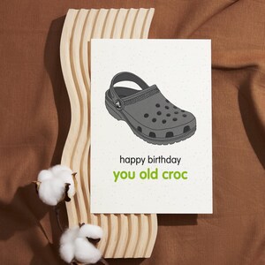 Puede incluir: Una tarjeta de cumpleaños con una ilustración de dibujos animados de un zapato de zueco gris. La tarjeta dice "feliz cumpleaños viejo cocodrilo".