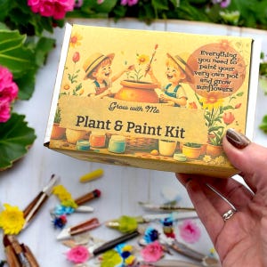 May include: A yellow box with a colourful illustration of two children painting a pot with sunflowers. The box says "Grow with Me Plant & Paint Kit" and "Everything you need to paint your very own pot and grow sunflowers".