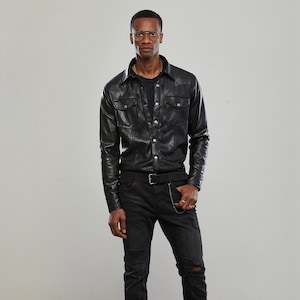 Könnte beinhalten: Ein schwarzes Lederhemd mit Knopfleiste und langen Ärmeln. Das Hemd hat zwei Brusttaschen und eine silberne Kette, die am Saum hängt. Das Model trägt eine schwarze Jeans mit Destroyed-Details und schwarze Chelsea-Boots.