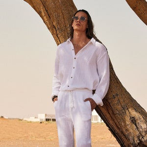 Peut inclure: Une chemise en lin blanche &agrave; manches longues et boutons en bois, associ&eacute;e &agrave; un pantalon blanc &agrave; cordon de serrage assorti. La tenue est port&eacute;e avec des sandales. La personne est appuy&eacute;e contre un arbre dans un paysage d&eacute;sertique.
