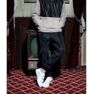 May include: A man wearing a black and beige fleece jacket with a zipper, black pants, and white sneakers with velcro straps. He is standing in front of a vintage television set with a dark wood frame.