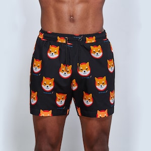May include: Black swim trunks with a repeating pattern of orange Shiba Inu dog faces. The dog faces have black eyes, red noses, and white teeth.