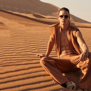May include: A person wearing a brown sleeveless vest, matching pants, and sunglasses sits in the desert. The outfit is a solid color. The background is a desert landscape with sand dunes.