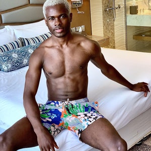 Puede incluir: Un hombre con el pelo corto y rubio platinado está sentado en una cama blanca con pantalones de baño de estampado colorido. Está sin camisa y tiene un físico musculoso.