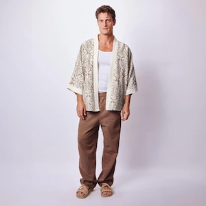 Puede incluir: Una camiseta sin mangas blanca combinada con pantalones marrones y un kimono de color crema abierto por delante con un patr&oacute;n geom&eacute;trico marr&oacute;n. El conjunto se completa con sandalias beige, creando un look en capas con tonos neutros y formas.