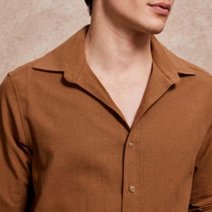 May include: A close-up of a brown button-down shirt with a wide collar. The shirt is made of a textured fabric and has three visible buttons. The color is a warm, earthy tone.