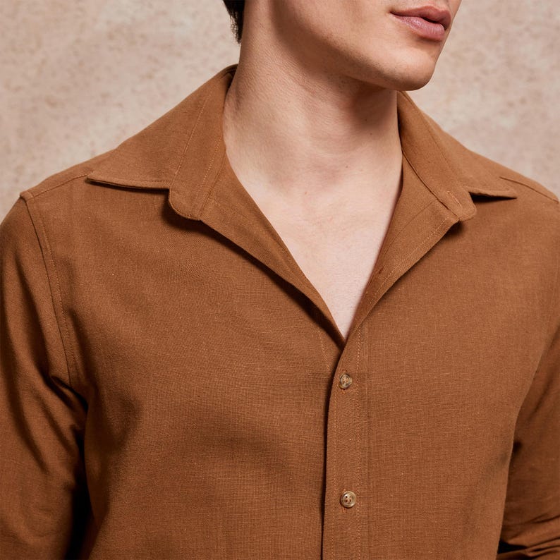 May include: A close-up of a brown button-down shirt with a wide collar. The shirt is made of a textured fabric and has three visible buttons. The color is a warm, earthy tone.