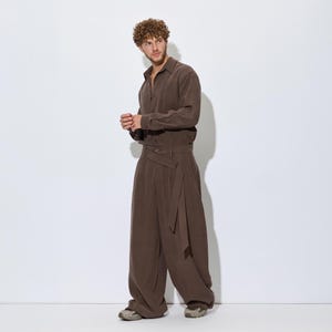 Op de afbeelding: Een bruin overhemd met lange mouwen en een broek met wijde pijpen. Het overhemd heeft een button-down kraag. De broek heeft een wikkel taille met een riem. De outfit is gemaakt van een zachte, vloeiende stof. Het model draagt sneakers.