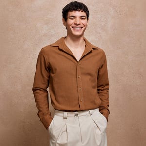 May include: A brown button-down shirt with a wide collar and long sleeves. The shirt is paired with cream-colored pleated pants. The model is smiling and looking at the camera.