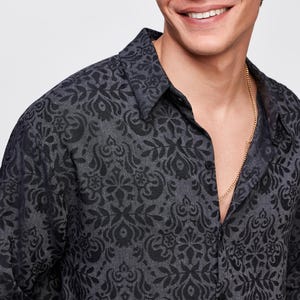 Op de afbeelding: Een donkergrijs overhemd met zwarte bloemenprint. Het overhemd heeft een klassieke kraag en is gedeeltelijk opengeknoopt, waardoor een gouden ketting zichtbaar is. De stof heeft een getextureerde uitstraling.