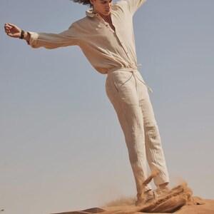 May include: A person wearing a beige linen shirt and matching pants, standing on a sand dune. The long-sleeved shirt is buttoned and the pants have a drawstring waist. The person is in motion, with arms outstretched against a clear blue sky.