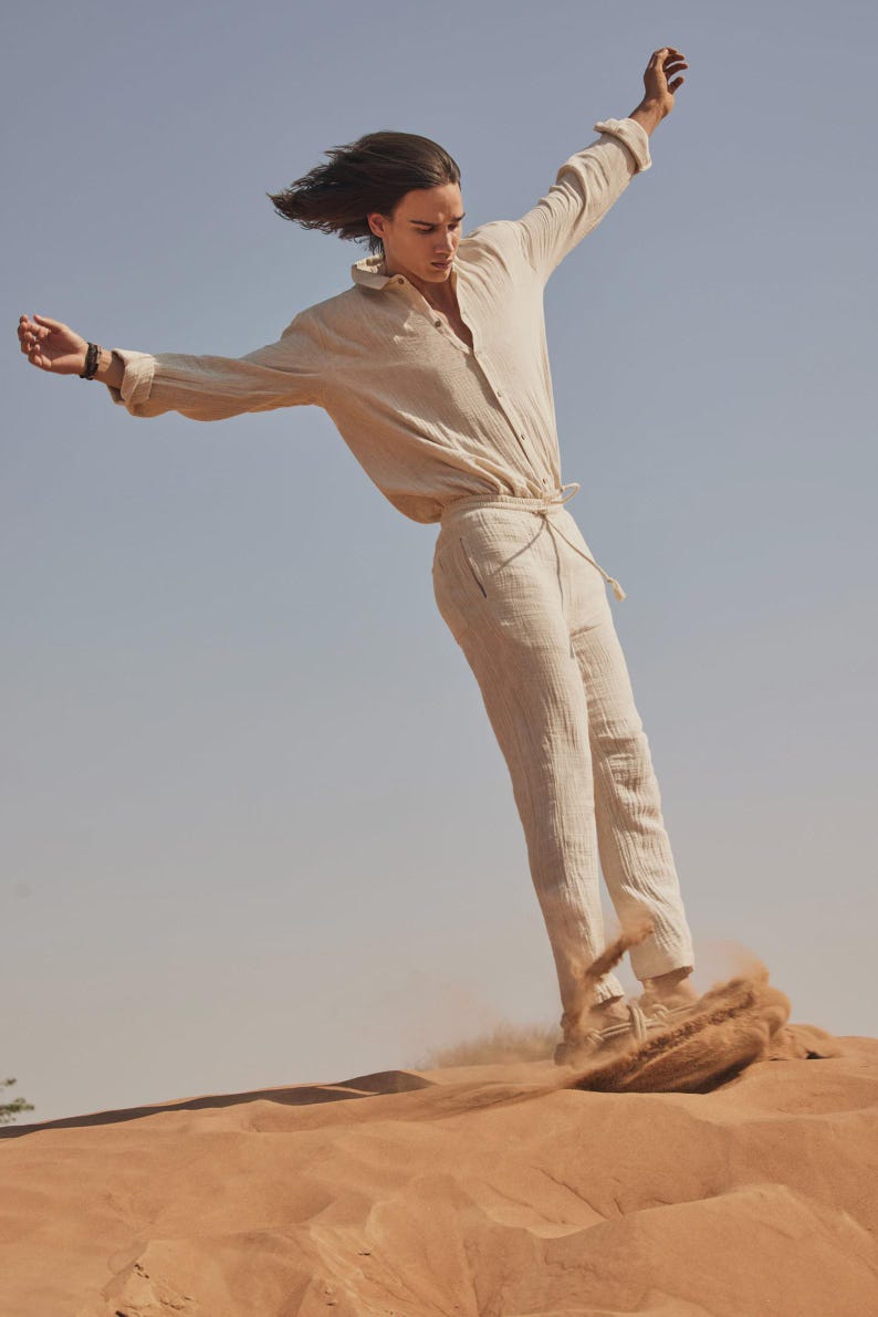 May include: A person wearing a beige linen shirt and matching pants, standing on a sand dune. The long-sleeved shirt is buttoned and the pants have a drawstring waist. The person is in motion, with arms outstretched against a clear blue sky.