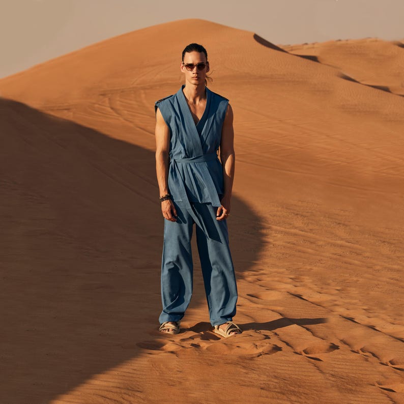 May include: A person in a blue sleeveless wrap top and matching trousers, posing in a desert setting. The outfit is belted at the waist. The person wears sunglasses and sandals. A large sand dune forms the backdrop.
