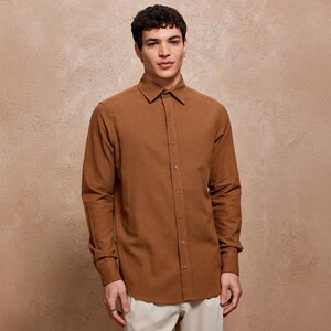 May include: A long-sleeved, button-down shirt in a warm brown hue. The shirt features a classic collar and buttoned cuffs. The fabric appears to have a subtle texture. The model is wearing the shirt with light-colored pants.