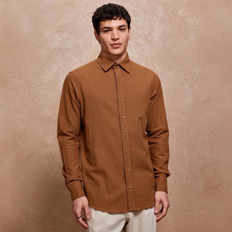 May include: A long-sleeved, button-down shirt in a warm brown hue. The shirt features a classic collar and buttoned cuffs. The fabric appears to have a subtle texture. The model is wearing the shirt with light-colored pants.