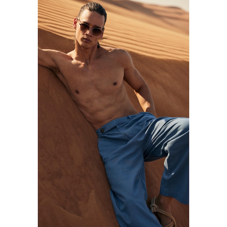May include: A person wearing blue trousers and sunglasses, leaning against a sand dune. The trousers have a button closure and a relaxed fit. The sunglasses have a brown frame. The background is a desert landscape.