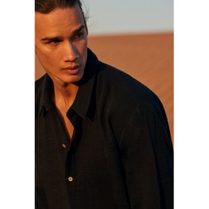 May include: A black button-down shirt with a classic collar and long sleeves. The shirt has two visible wooden buttons. The background is a desert landscape with warm, earthy tones.